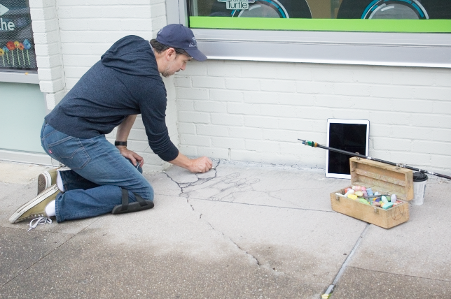 David Zinn bends down to create an anamorphic figure he’s become known for on a piece of cracked pavement in downtown Big Rapids.